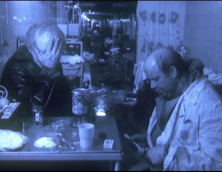 Two characters sitting by a metallic table. On the left, an old man hiding his face in his hands. On the right a balding white man. The scene is blue.