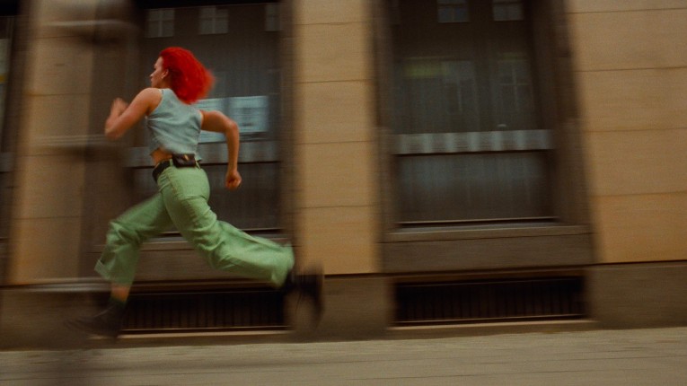 Red haied girl running on the left. A blurry building in the background